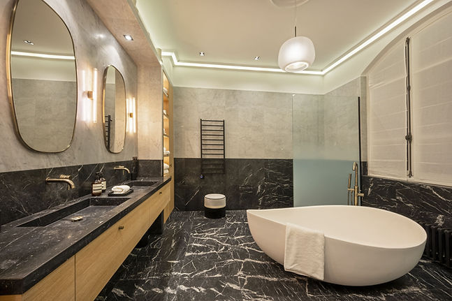 grande salle de bain au design moderne et luxueux. La décoration repose sur un fort contraste entre un sol et un bas de mur en marbre noir fortement veiné de blanc, et une partie haute des murs en carrelage gris clair. À gauche, un long meuble double vasque en bois est surmonté de marbre noir, de robinetteries dorées et de deux miroirs ovales avec éclairage mural. À droite se trouve une baignoire îlot blanche aux lignes organiques, accompagnée d'un robinet sur pied doré. L'arrière-plan révèle un radiateur porte-serviettes noir, un petit pouf rond et une paroi de douche en verre givré.