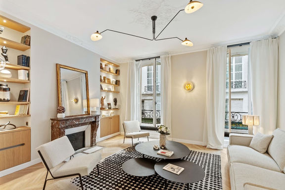 Un grand salon lumineux et élégant, mêlant architecture classique et mobilier contemporain. Au centre, trois tables basses rondes noires de tailles différentes s'imbriquent sur un grand tapis à motifs géométriques noirs et blancs. Sur la droite, un canapé en tissu texturé clair fait face à deux fauteuils blancs épurés à structure métallique noire. Sur le mur de gauche, une cheminée d'époque en marbre brun est surmontée d'un grand miroir rectangulaire à cadre doré. Elle est encadrée par deux niches aménagées sur mesure avec des étagères en bois clair illuminées, exposant des livres et des objets décoratifs, et des rangements fermés en partie basse. Au fond, deux grandes portes-fenêtres ouvertes, habillées de longs rideaux blancs, laissent entrer une belle lumière naturelle et offrent une vue sur des balcons parisiens en fer forgé. L'ensemble de la pièce est mis en valeur par un parquet à chevrons en bois clair au sol et des moulures décoratives au plafond, d'où est suspendu un grand luminaire noir moderne à plusieurs bras.