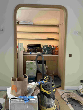 "Une vue de l'intérieur d'une maison en pleins travaux. Au premier plan, il y a du matériel de nettoyage et de bricolage : un gros aspirateur de chantier jaune et noir, des cartons et des sacs en papier posés sur un sol protégé. Au centre de l'image, on voit un grand passage ouvert avec un cadre en bois clair, dont les coins en haut sont arrondis. À travers ce passage, on regarde dans une autre pièce où se trouve un grand meuble en bois avec des étagères éclairées par des lumières jaunes. Cette petite pièce est encombrée de sacs, de valises, d'outils et de boîtes de rangement vertes empilées par terre.
