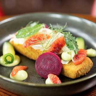 Gourmet dish with crispy patty, beet slice, citrus segments, green sauce blobs, and herbs on a dark plate. Vivid and artistic presentation.