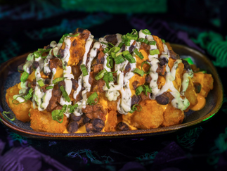 Loaded nachos topped with cheese, sour cream, black beans, and green onions on a dark plate. The background has a green and purple pattern.