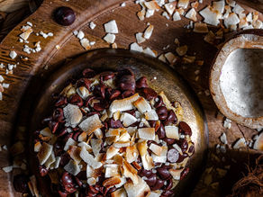 A wooden bowl filled with dark chocolate chips and coconut flakes on a wooden surface. Empty coconut shells nearby. Warm, rustic ambiance. This is the coconut caramel chocolate chip cookie available the first week of June at Gideon's Bakehouse.