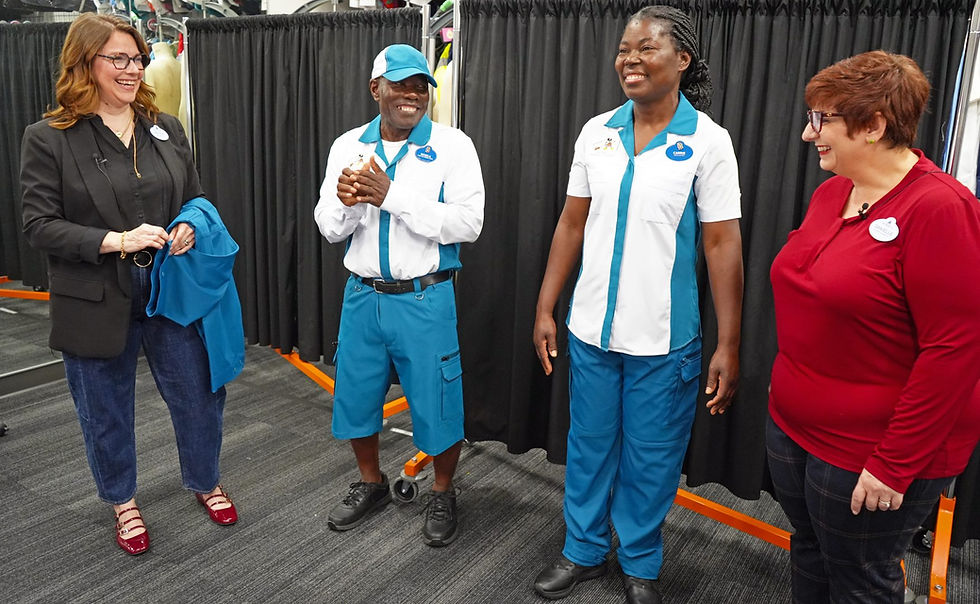 Four people smiling in a room with black curtains. Two wear blue and white uniforms, two in casual attire. Name tags visible. Casual, joyful mood.