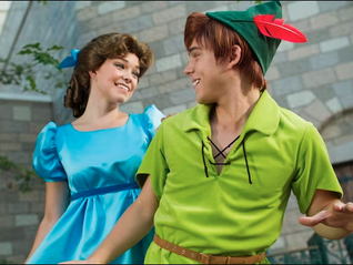 A person in a blue dress and another in a green outfit and hat smile at each other in front of a stone building, creating a playful mood.  These are Disney Characters Wendy Darling and Peter at Walt Disney World.