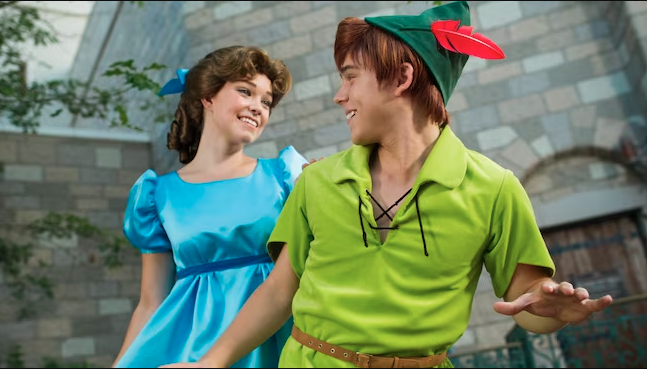 A person in a blue dress and another in a green outfit and hat smile at each other in front of a stone building, creating a playful mood. These are Disney Characters Wendy Darling and Peter at Walt Disney World.
