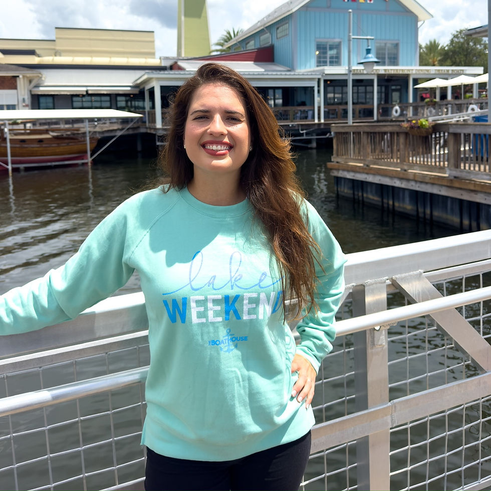 Smiling woman in a light blue "Lake Weekend" sweatshirt poses by a waterfront railing. Blue building and wooden deck in the background.