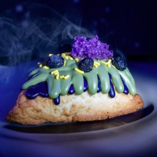 A scone with green and purple icing, topped with blueberries and purple sprinkles. Smoke rises around it, set against a dark, misty background.  Grab your Mossberry Biscuit From The May 4th Star Wars Foodie Guide at Disney's Hollywood Studios.