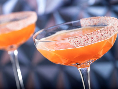 Two orange cocktails in glassware with sugared rims, set against a blurred geometric backdrop, creating a vibrant and festive mood.