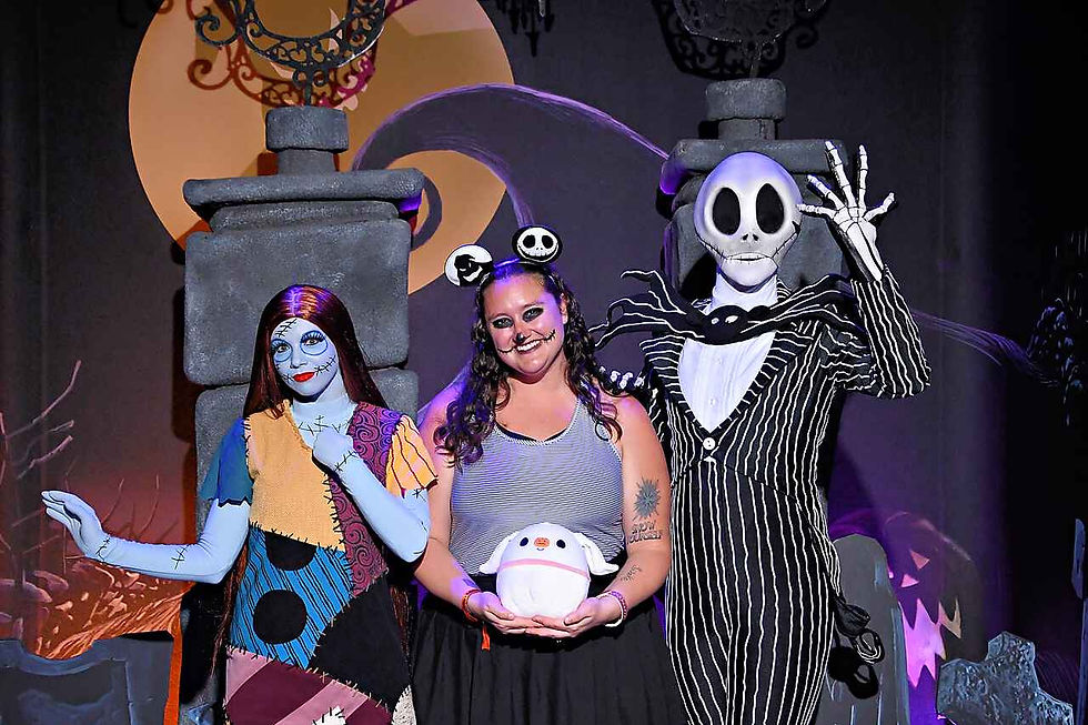 A woman poses with characters dressed in Halloween costumes, surrounded by spooky decorations and a large yellow moon background.  These are Jack Skellington and Sally character meets from The Nightmare Before Christmas.