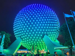 Illuminated geodesic sphere glows blue and green at night, surrounded by flags and trees. People gather below, creating a vibrant scene.