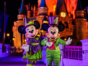 Mickey and Minnie Mouse in Halloween costumes pose cheerfully in front of a glowing castle at night, surrounded by bright lights.