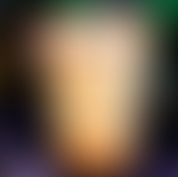 A glow-in-the-dark drink with a red sugar rim, orange syrup drips, and ice, sits on a Halloween-themed tablecloth. A pumpkin decoration is blurred in the background.