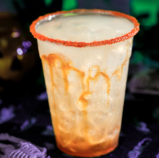 A glow-in-the-dark drink with a red sugar rim, orange syrup drips, and ice, sits on a Halloween-themed tablecloth. A pumpkin decoration is blurred in the background.