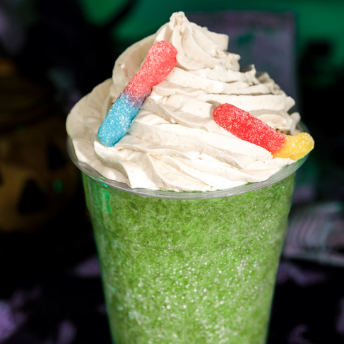A green slushie topped with whipped cream and gummy worms, set against a dark, spooky Halloween-themed background.