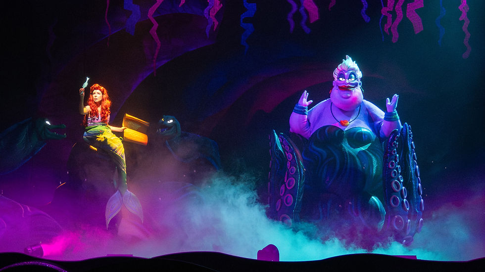 Stage scene from “The Little Mermaid: A Musical Adventure” featuring Ariel holding a fork while sitting on a rock with glowing yellow fish beside her, and Ursula towering nearby with outstretched tentacles, surrounded by colorful lighting and mist effects.