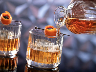 Crystal glass with amber liquid, ice, and an orange peel garnish. A decanter pours in front of a blurred geometric background.   This is the Brown Butter Old Fashioned cocktail at Geo 82 Lounge at Disney's Epcot.