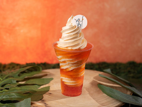 Rafiki Wildberry and DOLE Whip Orange Float. Swirled ice cream in an orange cup with cartoon character topper, set on wooden surface with green leaves. Warm, gradient background.