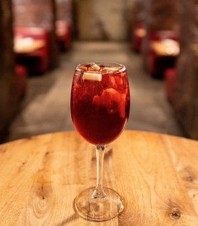 A glass of red sangria with fruit chunks sits on a wooden table in a dimly lit restaurant with red booths in the background.