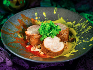 Rolled chicken with parsley garnish sits on colorful sauce splatters in a dark setting, with a small skeleton decoration nearby.