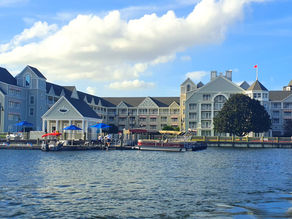Large, elegant waterfront hotel with red and blue umbrellas by the dock. Clear sky, calm water, and a relaxed atmosphere.