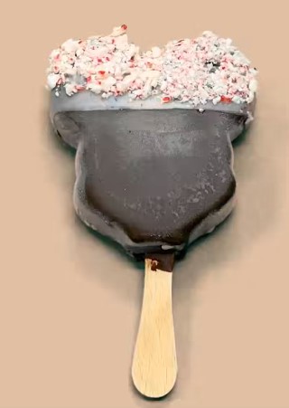 Heart-shaped chocolate ice cream with a wooden stick, topped with crushed white and red candy, on a light brown background.
