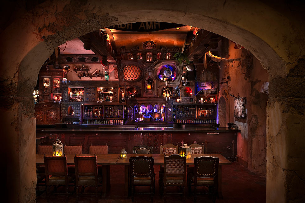 Dimly lit rustic bar with ornate wooden decor, flickering candles, and colorful lights. Empty chairs line a wooden table in the foreground.