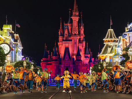 Colorful parade with dancers and a mascot under bright lights at a theme park. Castle backdrop glows red, creating a festive atmosphere.