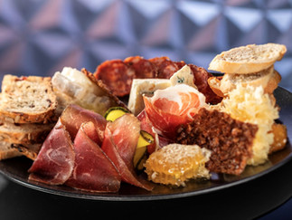 A platter of assorted cured meats, cheese, bread, and honeycomb on a black plate. Geometric pattern in the soft-focus background.   This is the Chef's Selection at Geo 82 Lounge at Disney's Epcot.