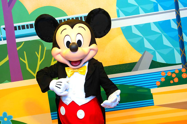 Mickey Mouse in red pants and yellow bow tie poses cheerfully against a colorful mural with geometric shapes and a blue train.