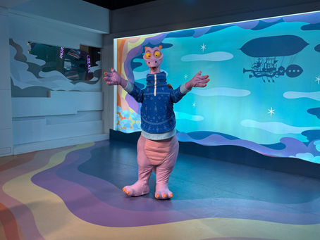 Figment in a blue sweater stands with open arms in a colorful room. Background shows a zeppelin and stars. Energetic mood.