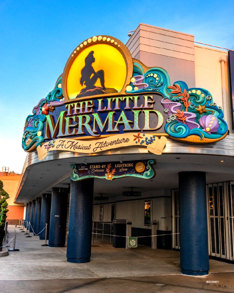 Colorful entrance to "The Little Mermaid: A Musical Adventure" with a whimsical sign featuring Ariel's silhouette against a yellow and orange sunset, surrounded by blue sea and purple shells. The setting is under a clear blue sky.