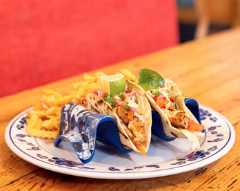 Fish tacos with fries on a floral plate, blue taco holder. Lime garnish. Set on a wooden table, red background adds warmth.