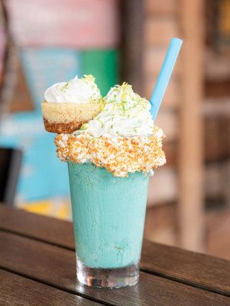 Blue milkshake in a glass with whipped cream and a mini cheesecake on top, blue straw. Set on a wooden table in a colorful cafe.