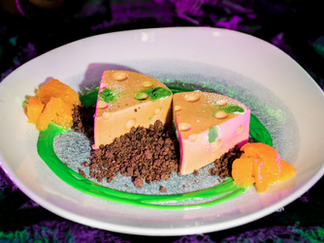 Two triangular orange mousse slices on a white plate with chocolate crumbles, green sauce, and orange segments, set on a colorful background.