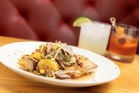 Plate of grilled fish with vegetables on a wooden table. Two cocktails behind on a red cushioned booth. Warm and inviting atmosphere.