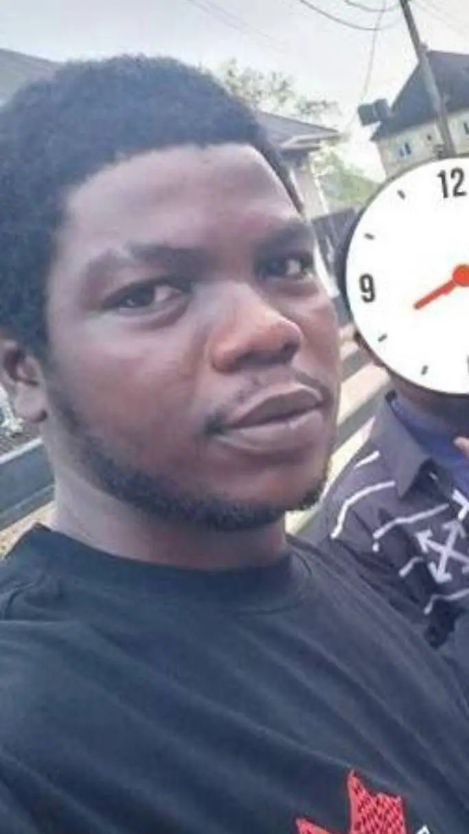 Man in a black shirt stands outdoors, holding a clock showing 9:00. Background includes trees and buildings. Neutral expression.