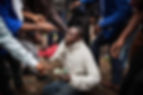 A man in a white sweater kneels and gestures intensely amid a crowd. People point at him, creating a tense atmosphere. Red glove visible.