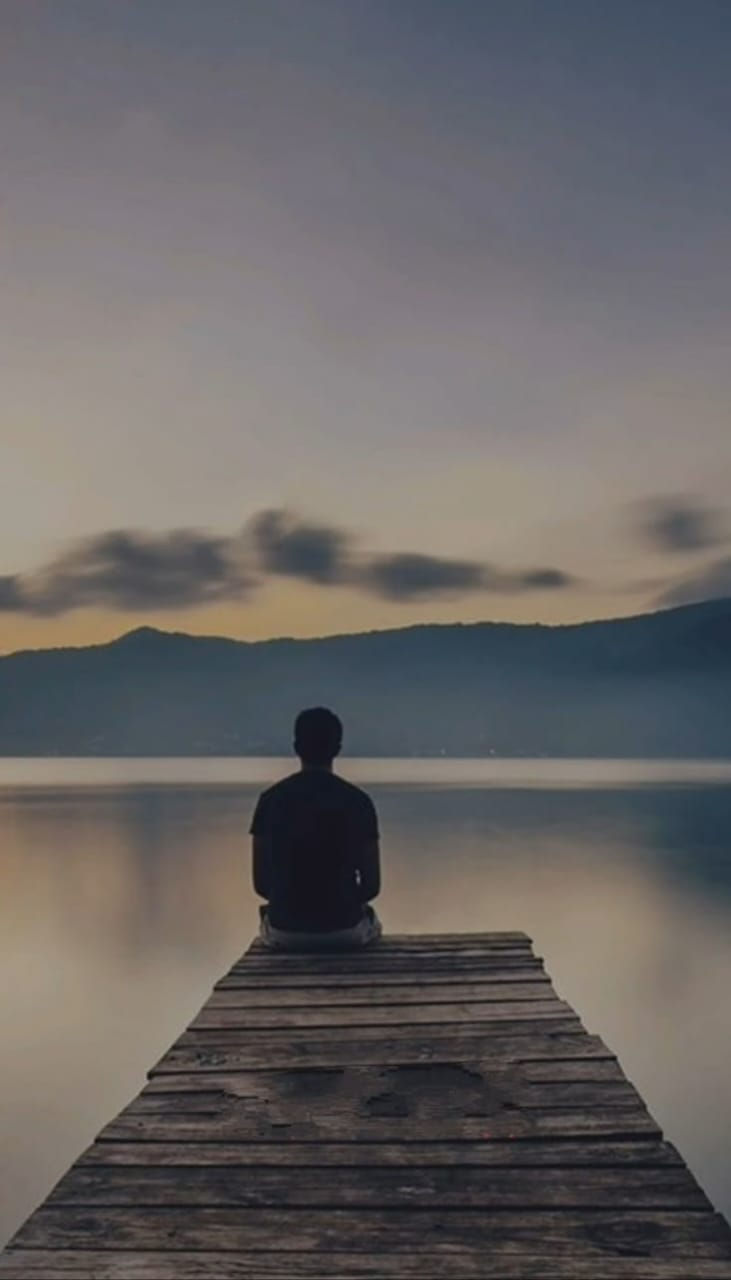 A person sits on a wooden dock, gazing at a serene lake during sunset. Silhouetted mountains and clouds enhance the tranquil mood.