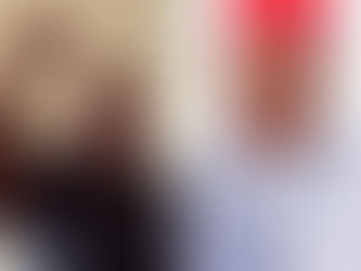 Woman raising fist in a room, wearing black. Man beside her in red cap and patterned attire. Neutral background. Confident mood.