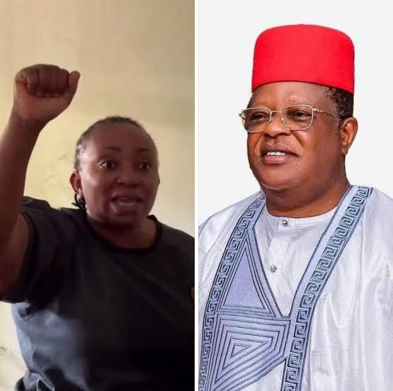 Woman raising fist in a room, wearing black. Man beside her in red cap and patterned attire. Neutral background. Confident mood.