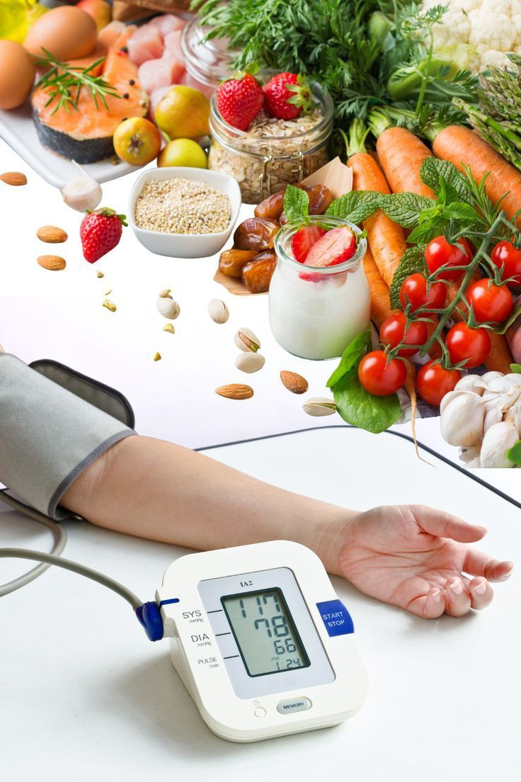 A blood pressure monitor reads 117/78 beside a person's arm. Fresh fruits, veggies, and grains are in the background, suggesting healthy eating.