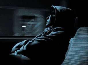 Person in a hoodie sits on a train seat, gazing out the window. Reflected in glass, serene mood, dim blue lighting, striped seat.