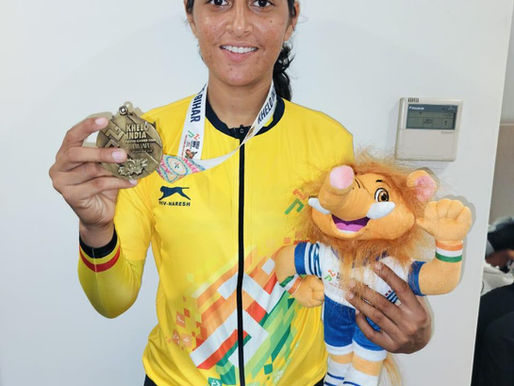 Manju Choudhary wearing a yellow cycling jersey, holding her Khelo India gold medal in one hand and a lion mascot in the other, smiling after her victory in the 20km Individual Time Trial event.
