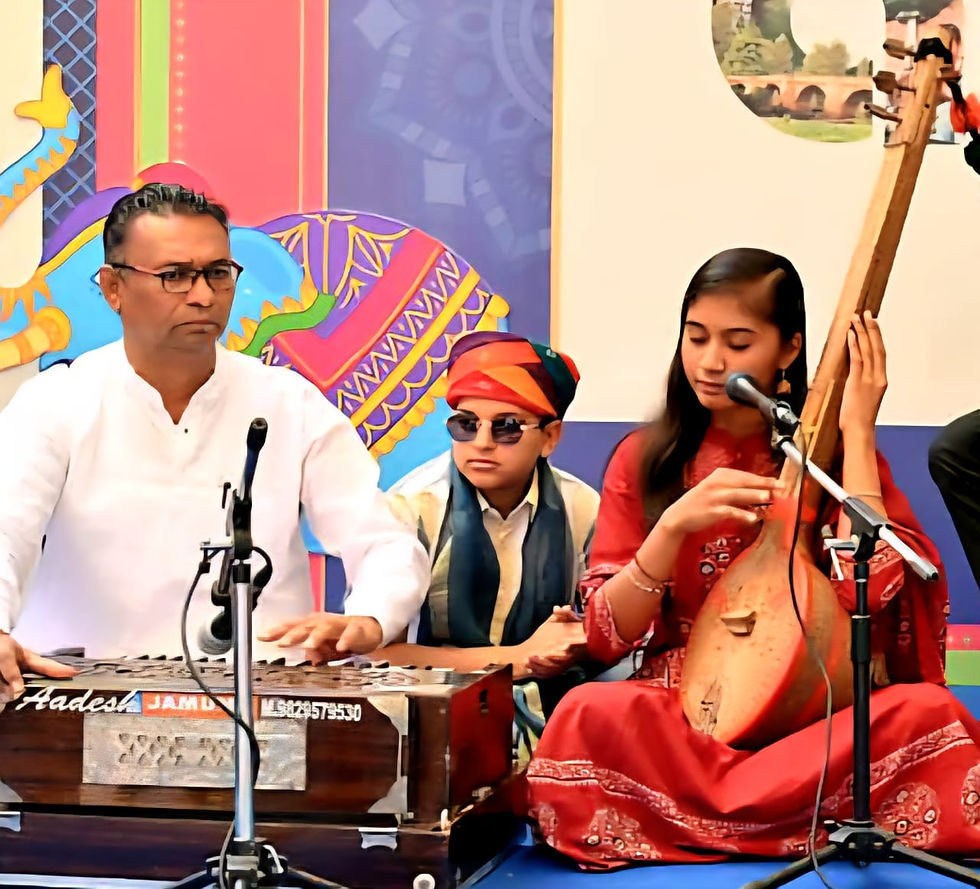 A man plays a harmonium and a woman strums a stringed instrument. A young boy in sunglasses watches. Colorful backdrop with patterns.