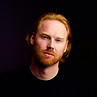 Red haired, bearded man in black t shirt looking at camera
