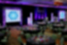 Business meeting event setup with professional lighting, projection screens, stage décor, speaker podium, and elegant, empty table seating.