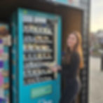 A smiling young woman in black clothing stands next to a brightly lit, turquoise fresh food vending machine. The machine, labeled 'Tuckshop,' is full of individually packaged fresh food, sweet treats, and snack food items. The woman is holding a freshly dispensed, pre-packaged item in her hands. Keywords: fresh food vending machine, sweet treats vending machine, snack food vending machines, Snacks food, fresh cookies 