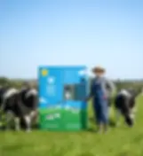 A wooden vending hut adorned with blue and white bunting contains two brightly colored "Whole Moo World" milk vending machines (one blue, one green) and a blue "Tuckshop" snack machine. A sign states the location is "Open 24/7.