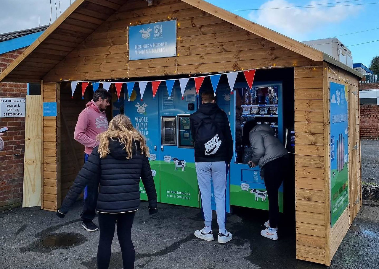 People using a 24 hour milk vending machine inside a small wooden shed, selling fresh milk and milkshakes