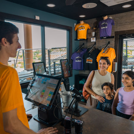 A smiling employee in an orange shirt helps a mother with two children at a store counter. T-shirts and roller coaster are visible in the background.